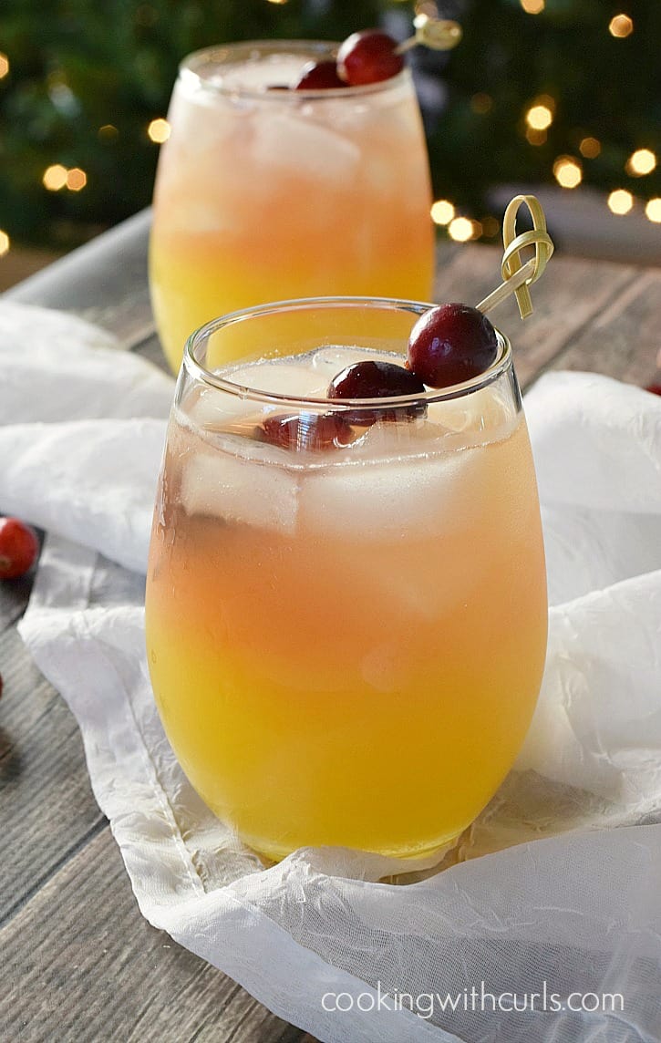 two clear glasses filled with orange, pineapple and cranberry juice garnished with fresh cranberries on a bamboo stick with a lit Christmas tree in the background.