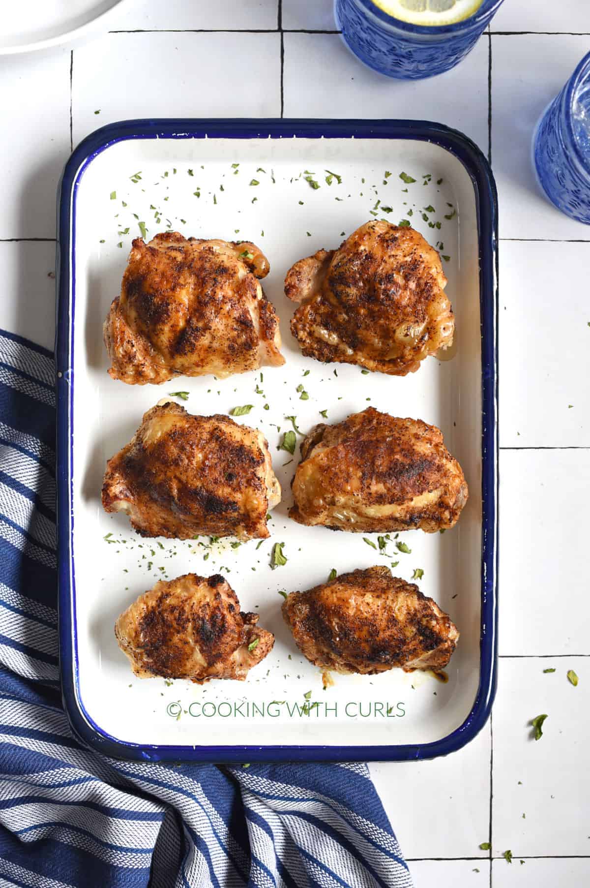 Six crispy bone-in chicken thighs on a white platter with dark blue rim, sitting on a blue and white striped towel with two blue glasses in an right hand corner. 