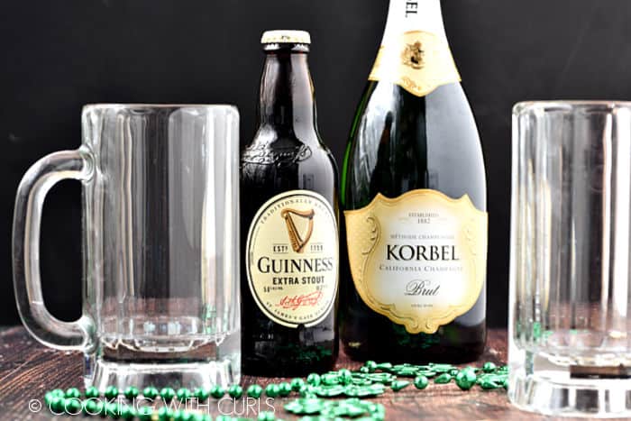 A bottle of Guinness beer, a bottle of Korbel champagne and two empty beer tankards with green beads on a wooden surface. 