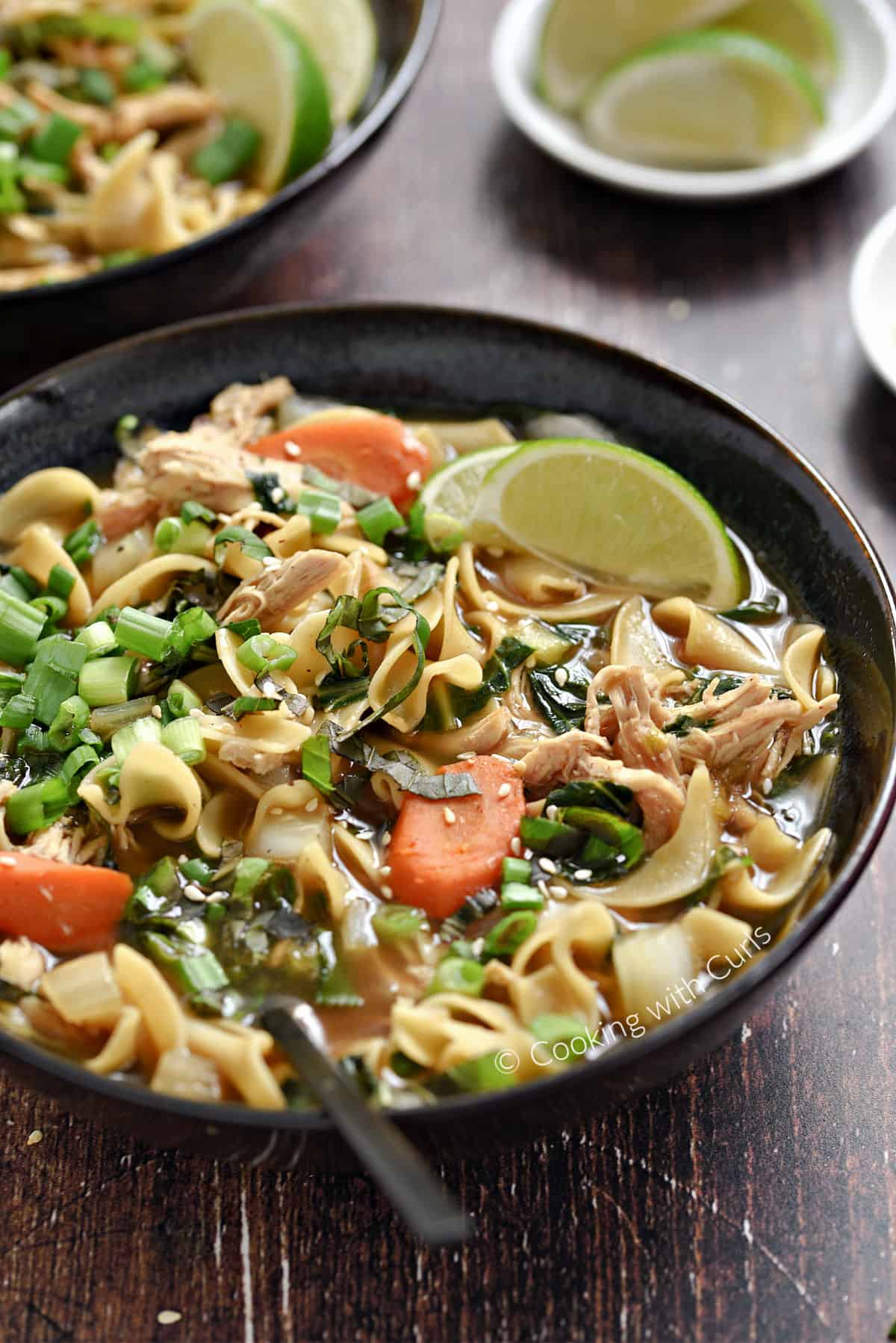 A bowl of Chinese Chicken Noodle Soup with carrots, green onions, and lime wedges with a second bowl of soup and lime wedges in the background. 