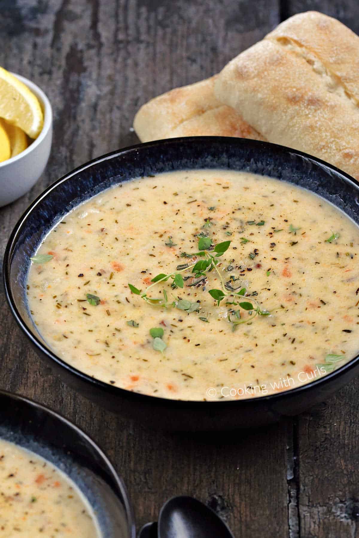 A bowl of creamy clam chowder with French rolls and a small bowl of lemon wedges in the background. 