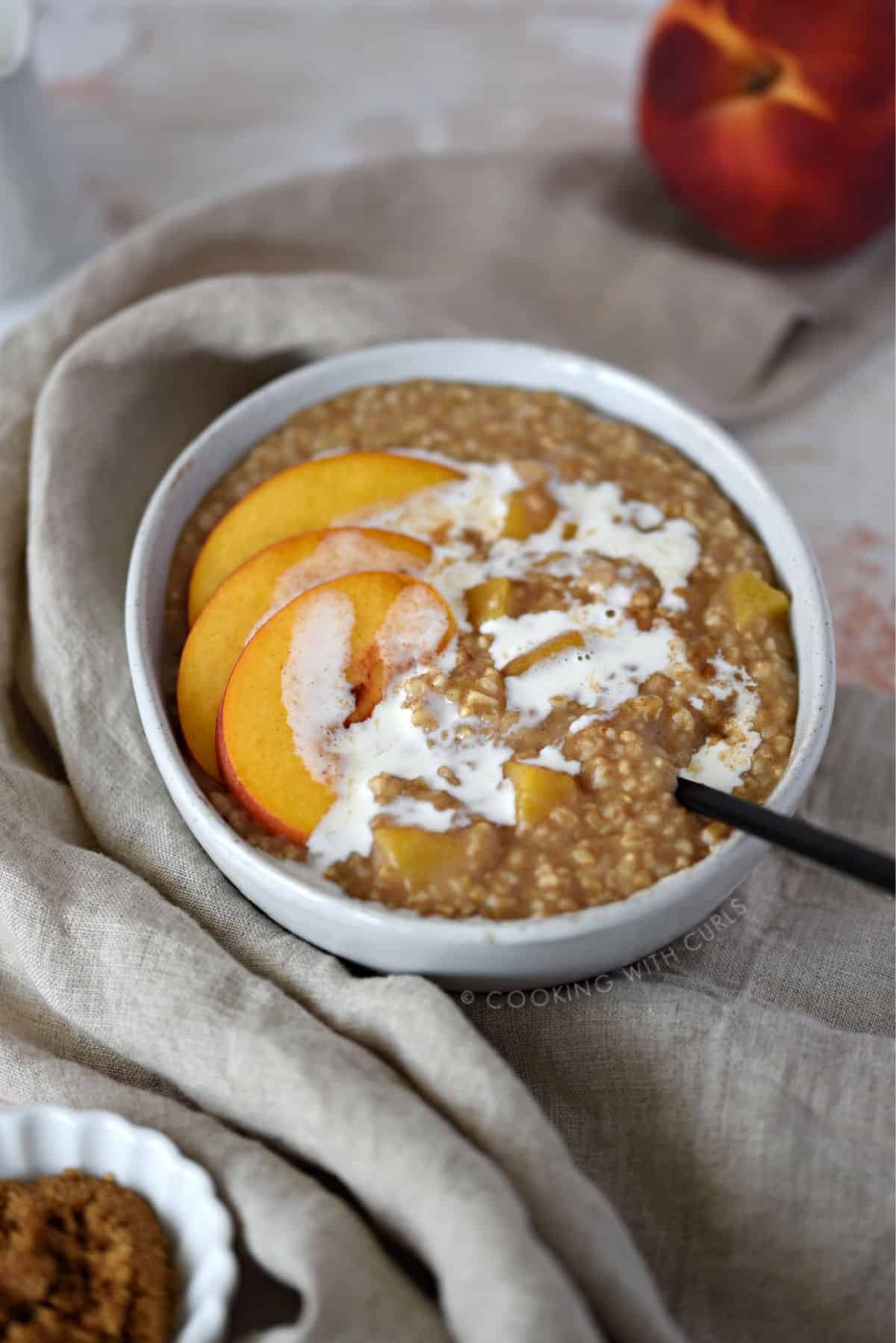 a big bowl of steel cut oats and chopped peaches topped with peach slices and heavy cream wrapped in a tan napkin with a black spoon sticking out of the bottom right side.