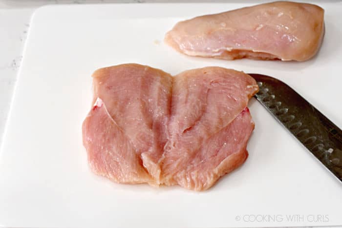 a butterflied chicken breast laying on a white cutting mat next to a chefs knife and second piece of chicken. 