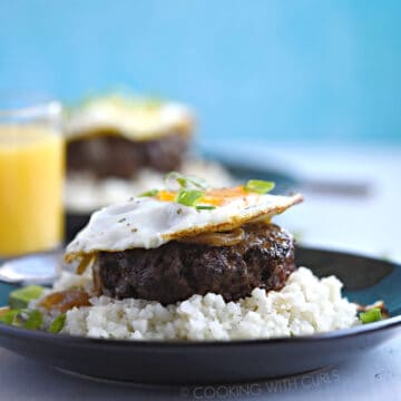 A ground hamburger steak patty topped with rich gravy and fried egg sits atop steamed rice and is garnished with green onions with a glass of orange juice and a second plate of food in the background.