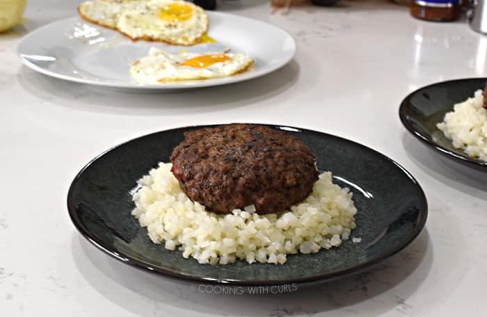 a cooked burger patty on a bed of rice with a plate of fried eggs in the background. 