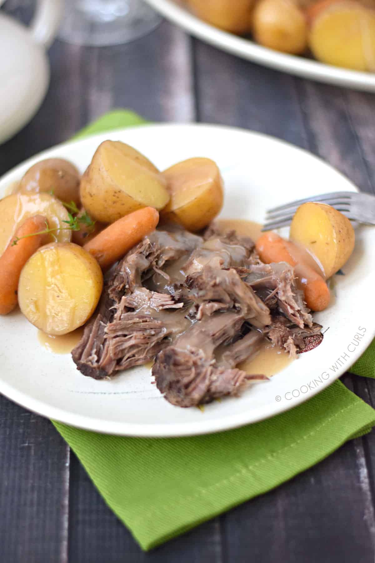 pot roast with sliced potatoes and baby carrots topped with beef gravy on a white plate that is sitting on a green napkin.