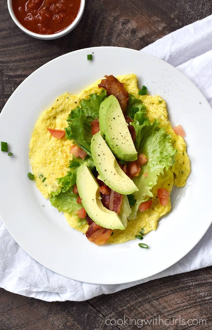 Three slices of avocado, two slices of bacon, lettuce leaves and chopped tomato laying on a flat egg "tortilla".