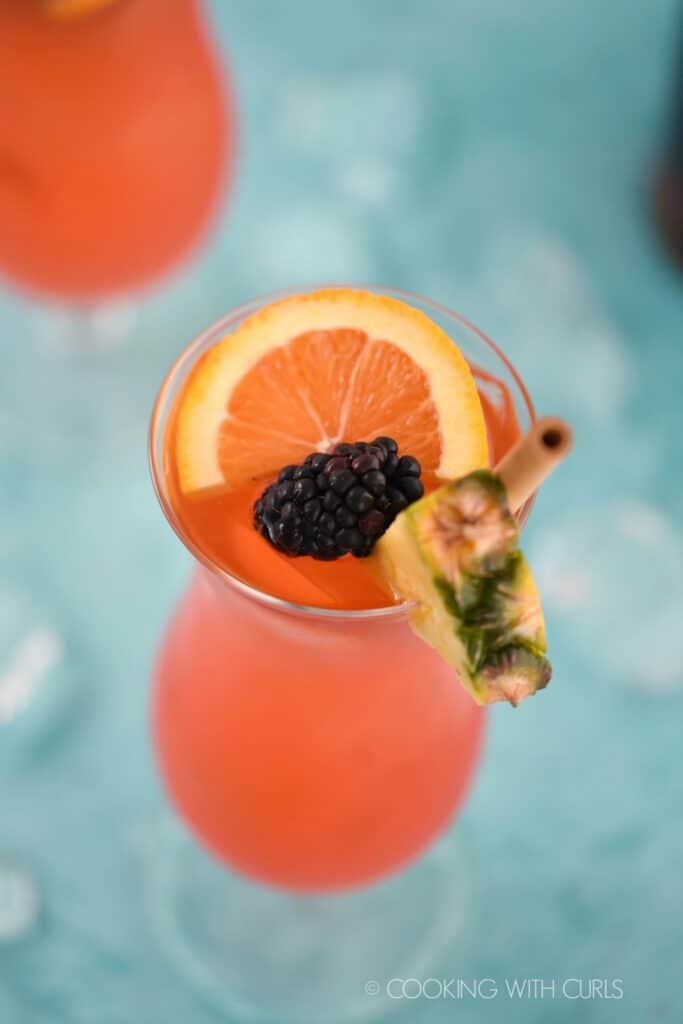 looking down on a pink cocktail garnished with a bamboo straw, pineapple wedge, orange slice and blackberry with a second cocktail in the upper right corner.