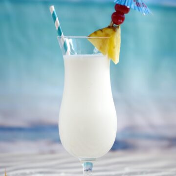 Creamy Pina Colada Milkshake in a hurricane glass garnished with a pineapple wedge, two cherries and a blue umbrella.