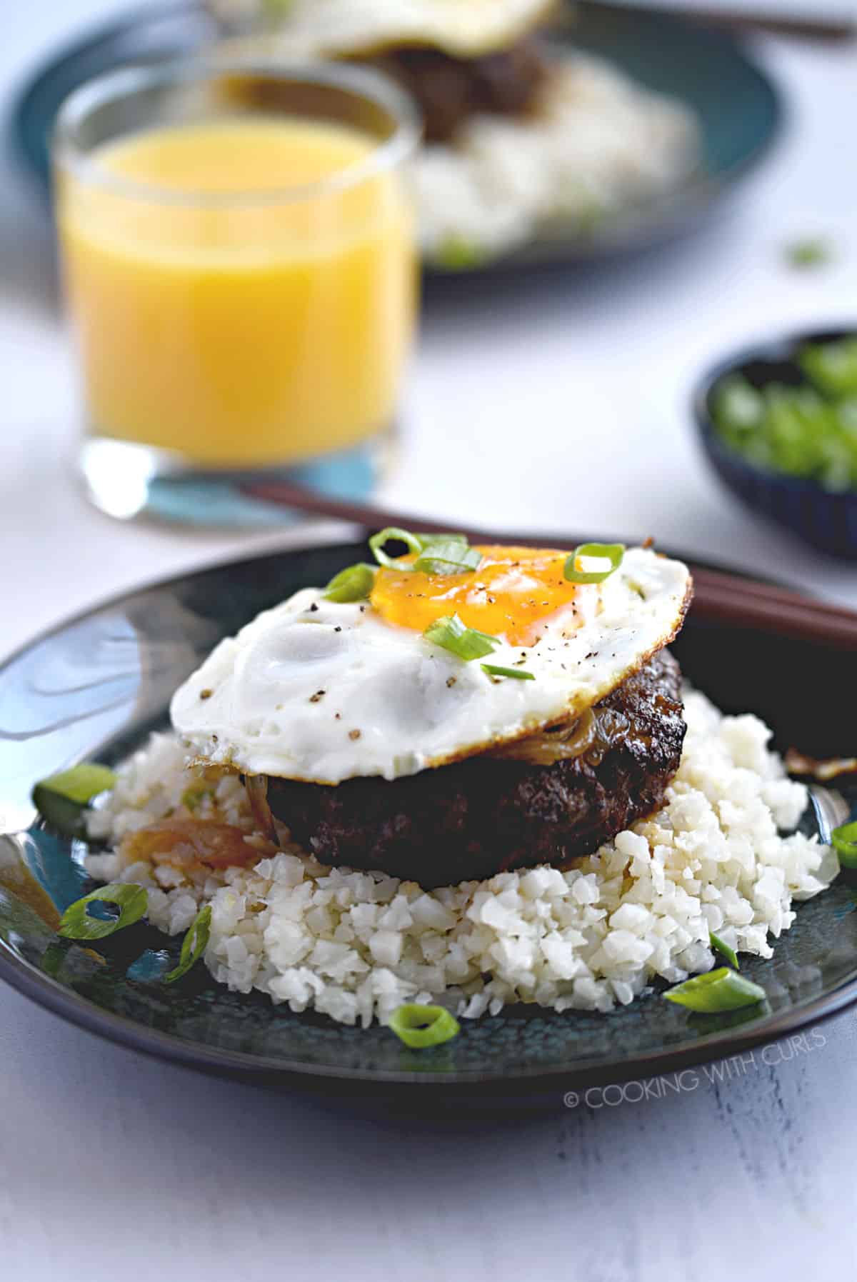a burger patty topped with brown gravy, fried egg and green onions on a bed of rice with a glass of orange juice and a second plate of food in the background.
