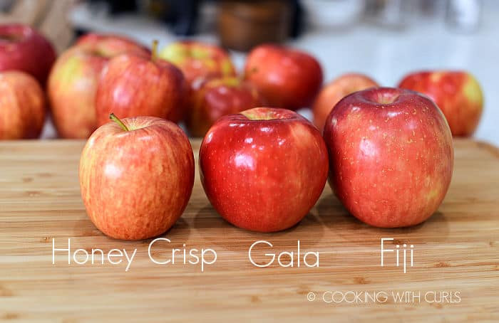 a honey crisp, gala, and fuji apple lined up on a wood cutting board with more apples in the background. 