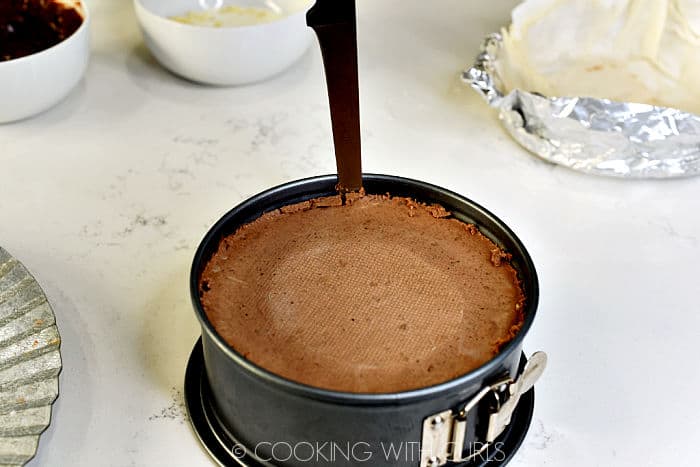 A knife running along the edge of the cheesecake and springform pan with bowls of chocolate in the background. 