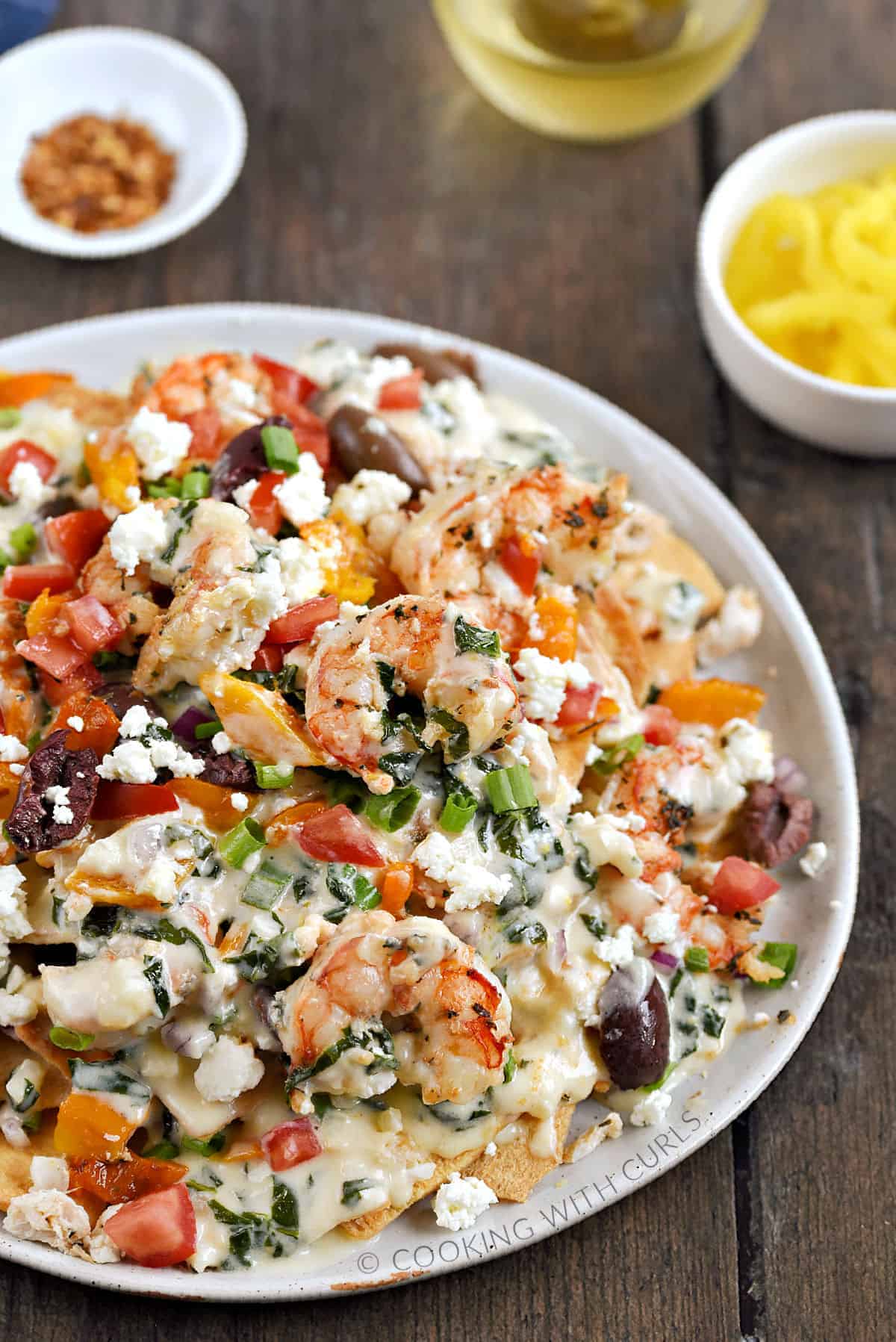 A large plate topped with shrimp, crab, diced tomatoes, olives, orange peppers, green onions, pita chips, and spinach cheese sauce. 
