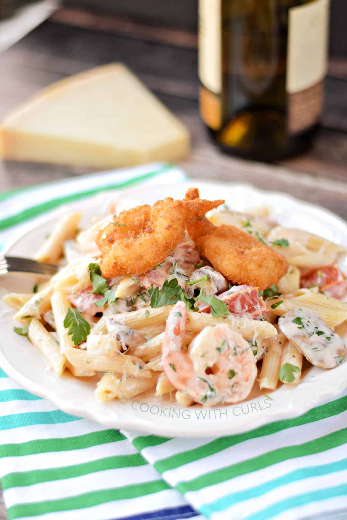 Penne pasta mixed with a creamy shrimp sauce, shrimp, mushrooms, tomatoes, and topped with three breaded shrimp on a white plate.