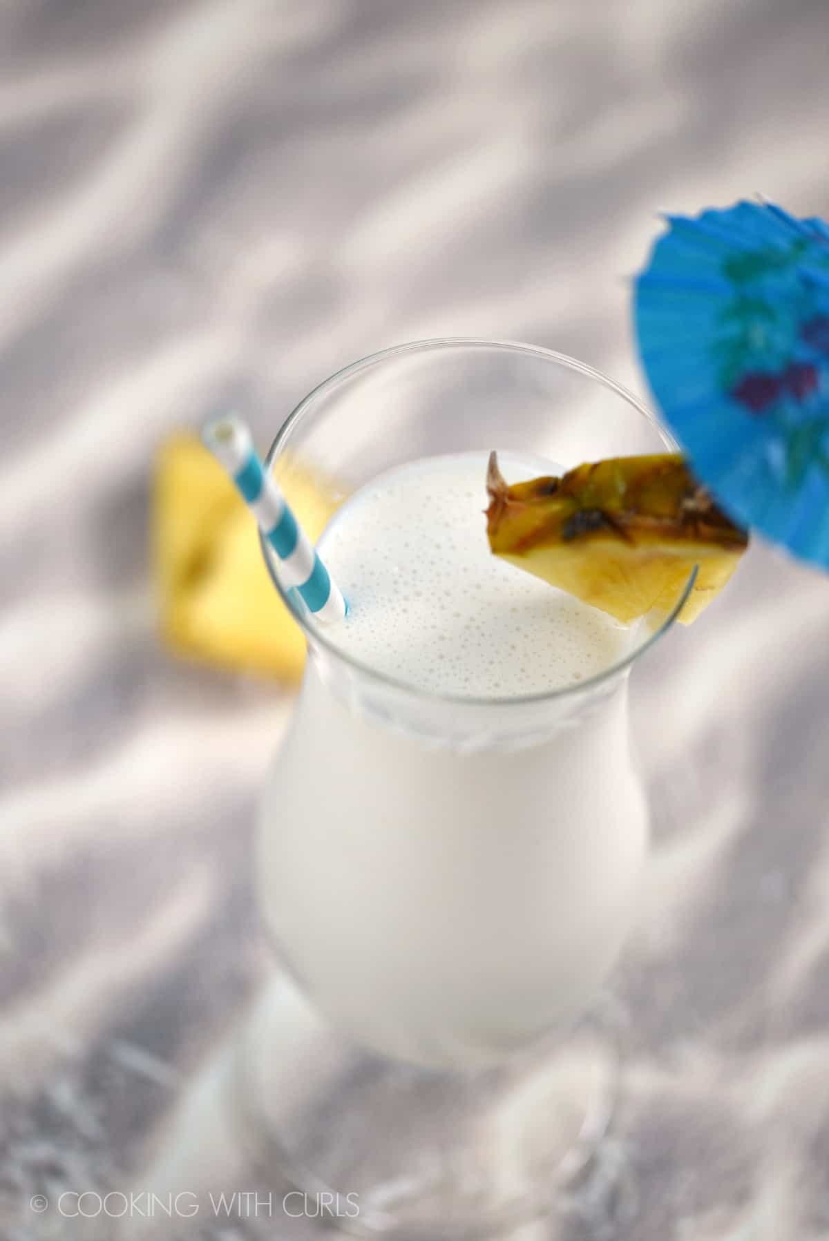 Looking down on a Pina Colada Milkshake in a tall glass garnished with a pineapple wedge, paper umbrella and striped straw.