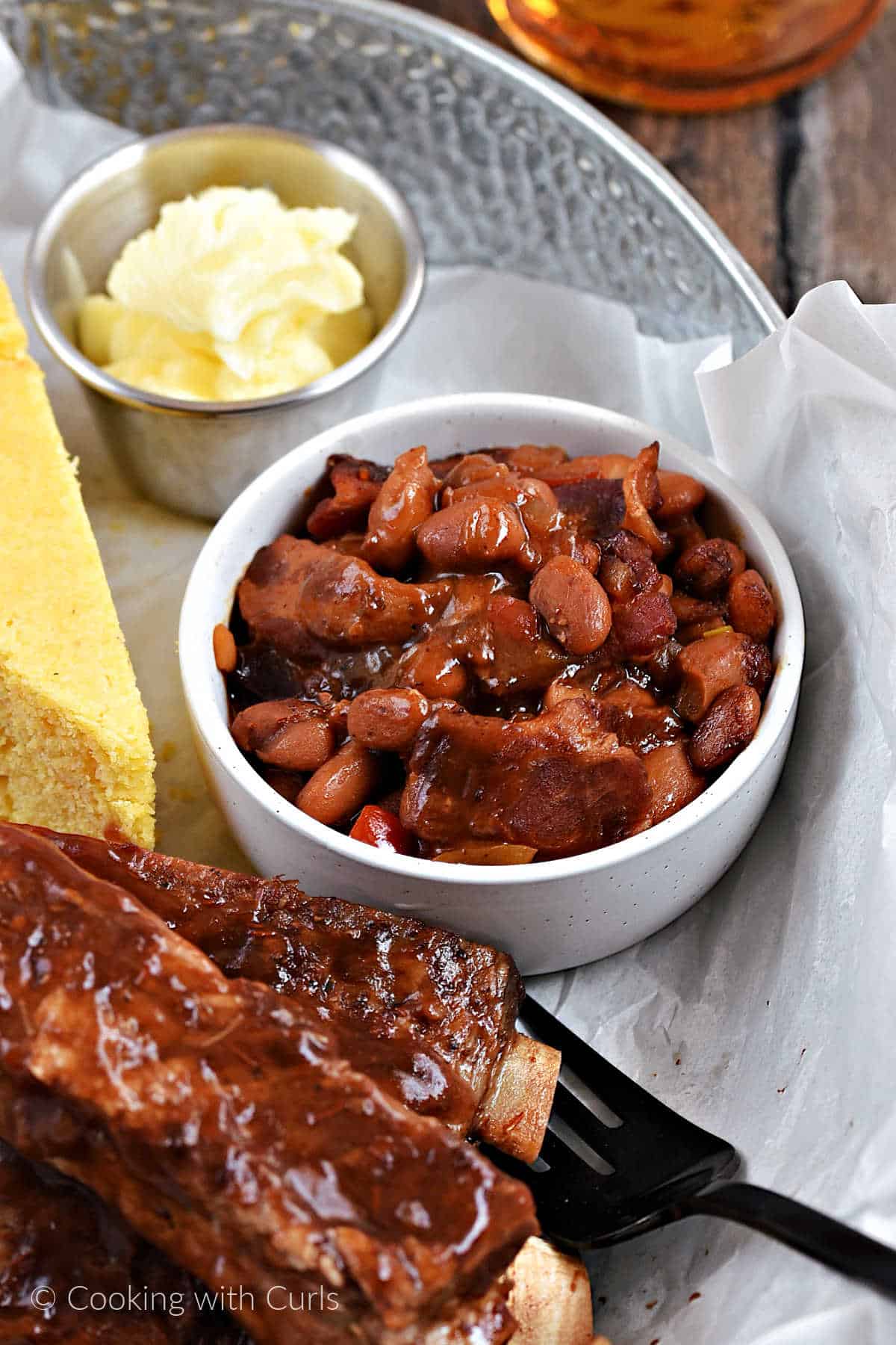 A small bowl of baked beans with chunks of bacon in a tray with barbecue ribs and cornbread.
