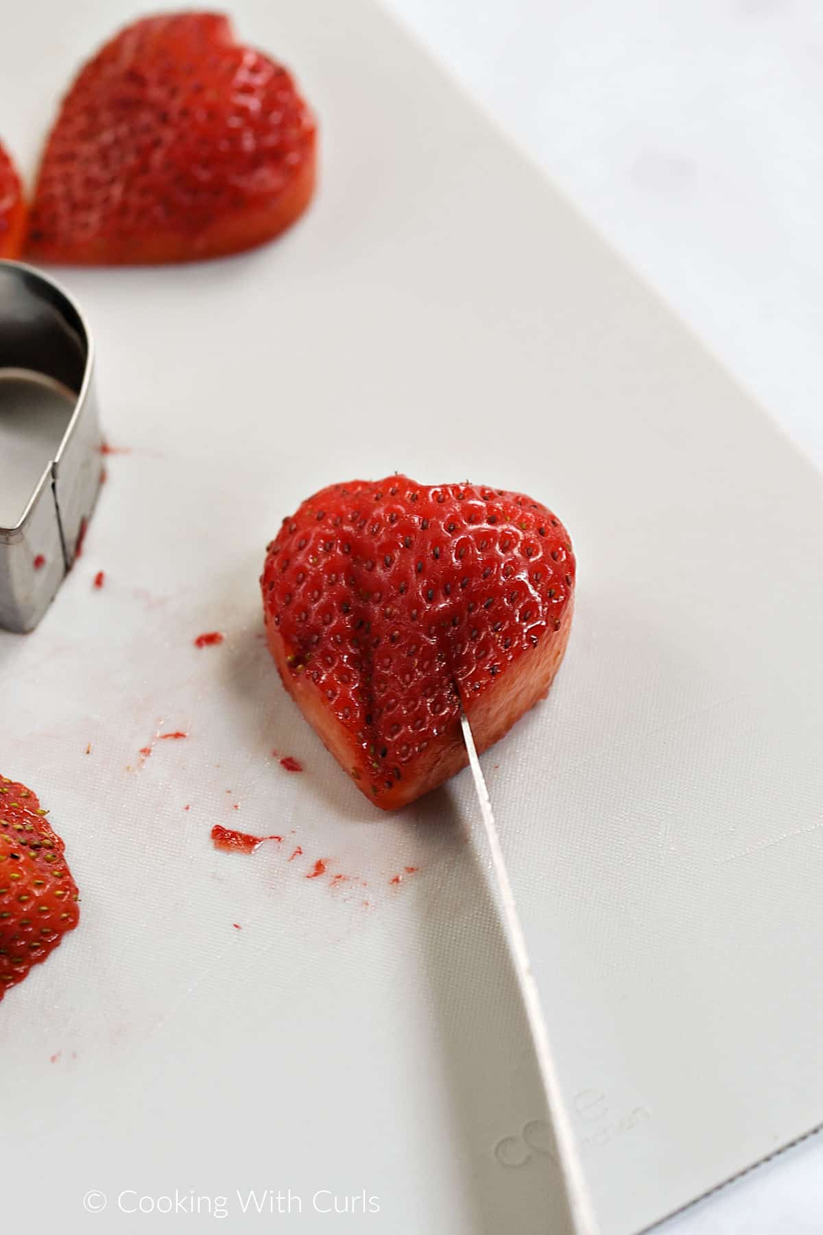 A small knife cutting a slit into the bottom corner of a heart shaped strawberry. 