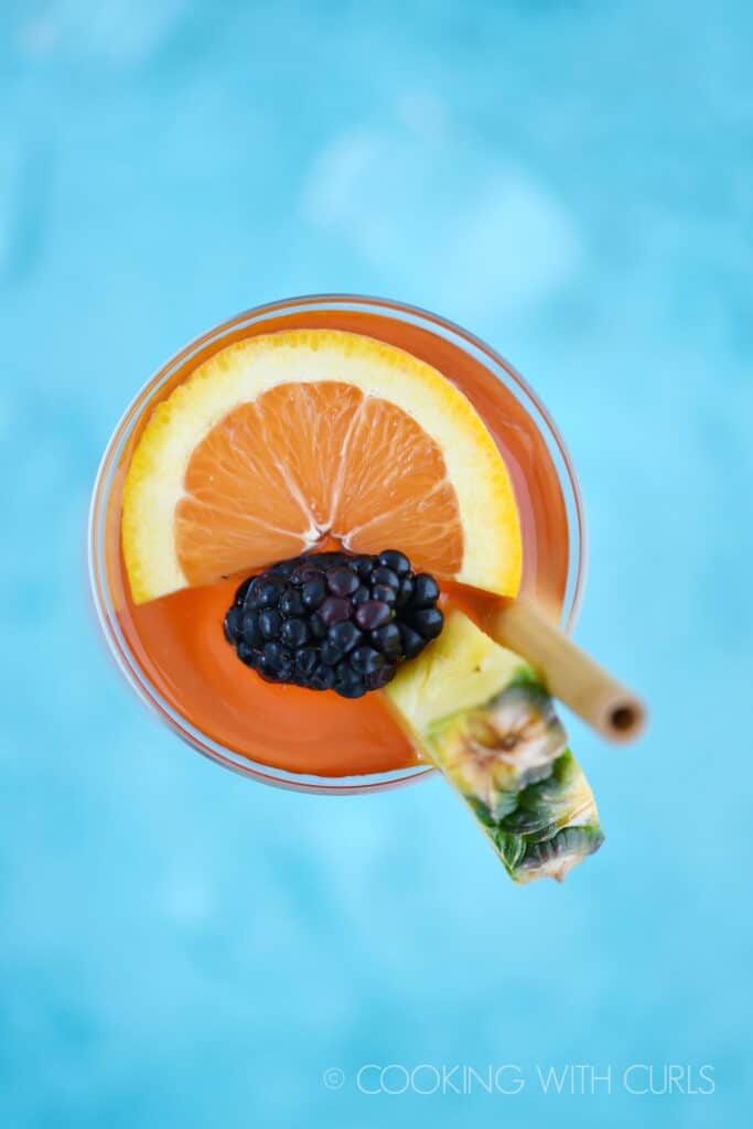looking directly down on a pink cocktail garnished with a pineapple wedge, orange slice, blackberry and bamboo straw sitting on a bright blue background.