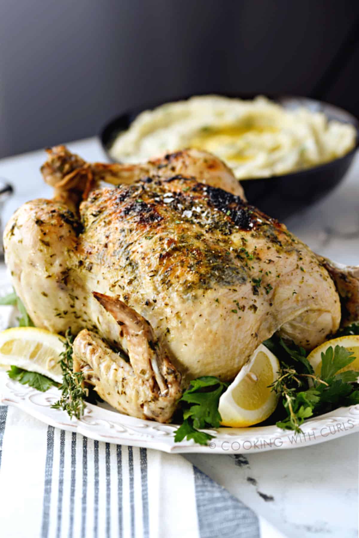 A whole Greek chicken on a white platter surrounded by fresh herbs and lemon wedges with a bowl of mashed potatoes in the background.