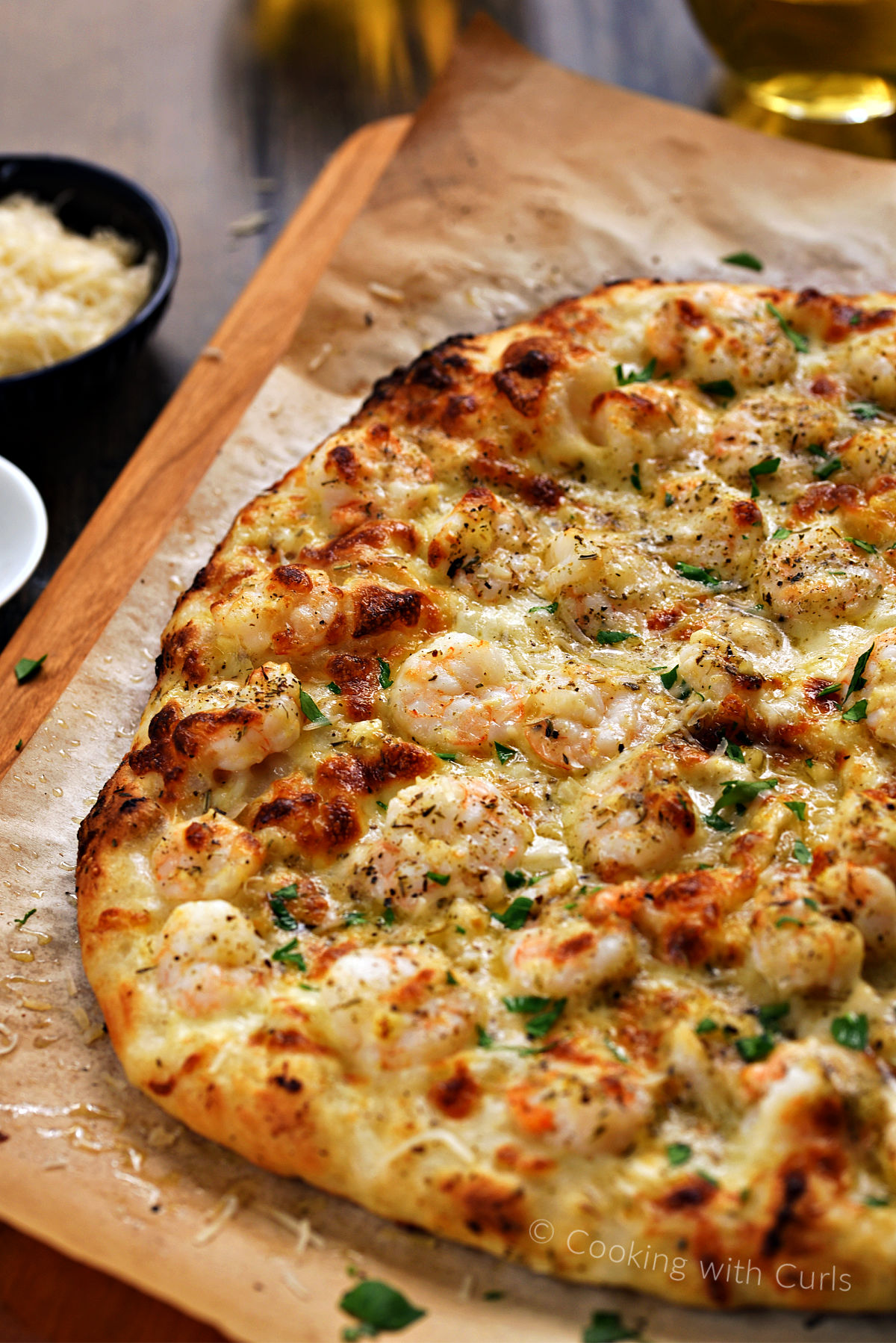 A whole shrimp scampi pizza on a wooden pizza peel with wine glasses in the background.