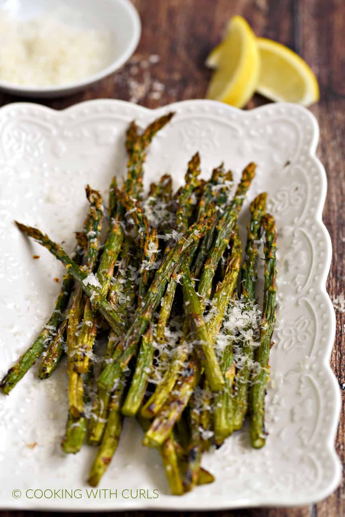 Asparagus spears topped with parmesan cheese and lemon zest on a white platter with a bowl of cheese and two lemon wedges in the background.