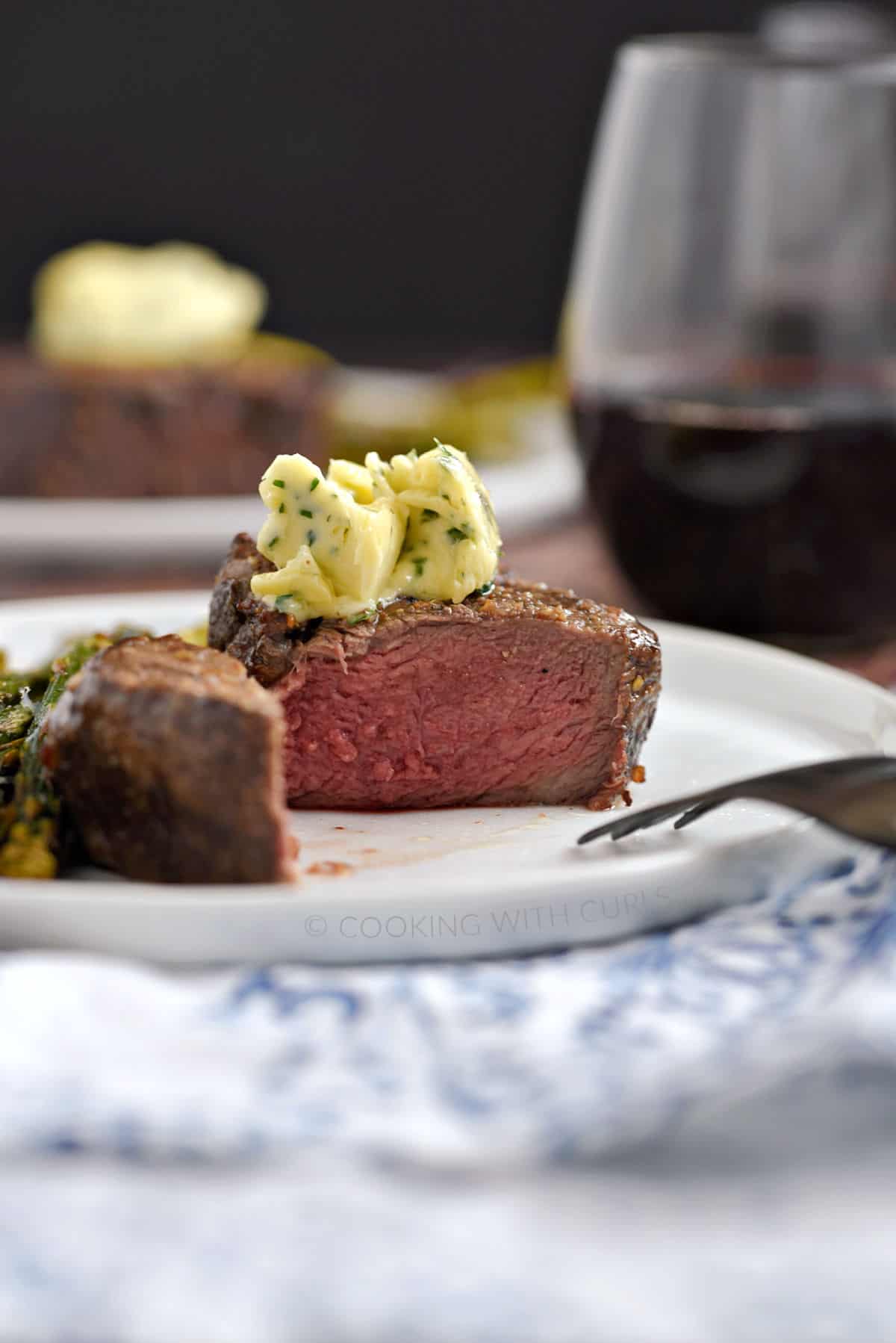 A filet mignon cut in half to show the pink center topped with herb butter on a white plate.