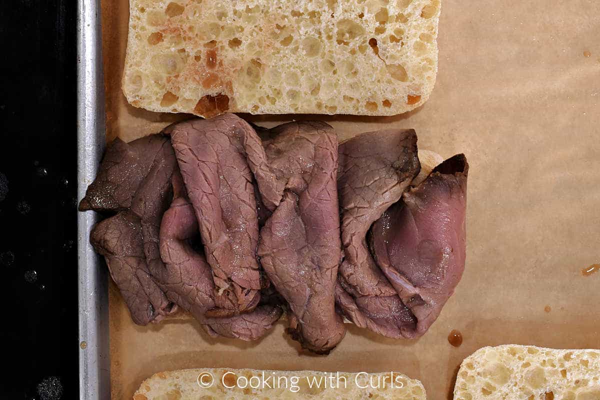 Au jus coated roast beef slices on a French roll sitting on a parchment lined baking sheet. 