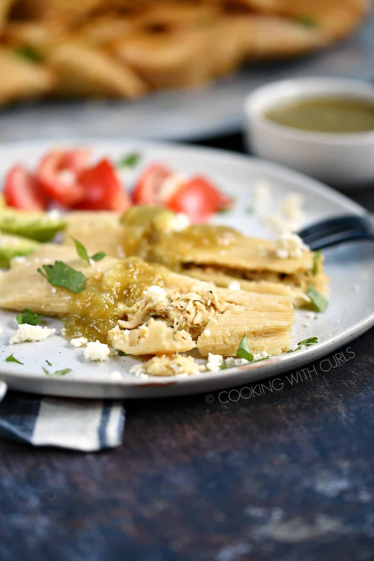 Two salsa verde chicken tamales on a white plate garnished with sliced avocado and tomatoes.