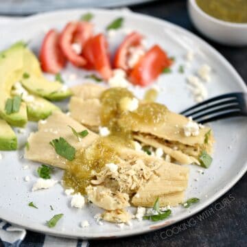 Two Instant Pot Salsa Verde Chicken Tamales drizzled with salsa on a white plate with sliced avocado and tomato in the background.