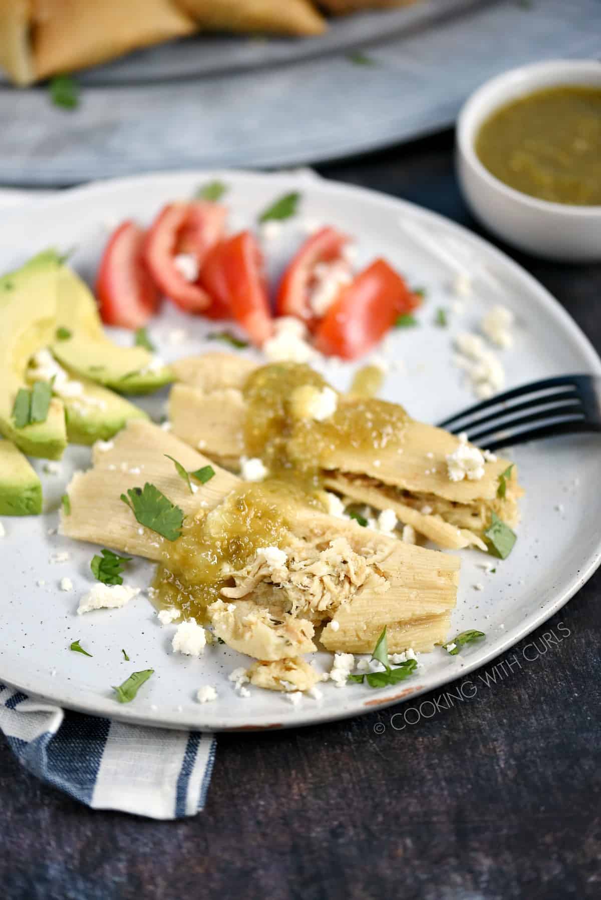 Two Instant Pot Salsa Verde Chicken Tamales drizzled with salsa on a white plate with sliced avocado and tomato in the background.