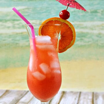 A bright pink Bahama Mama drink in a hurricane glass garnished with an orange wheel, cherry, and pink paper umbrella at the beach.