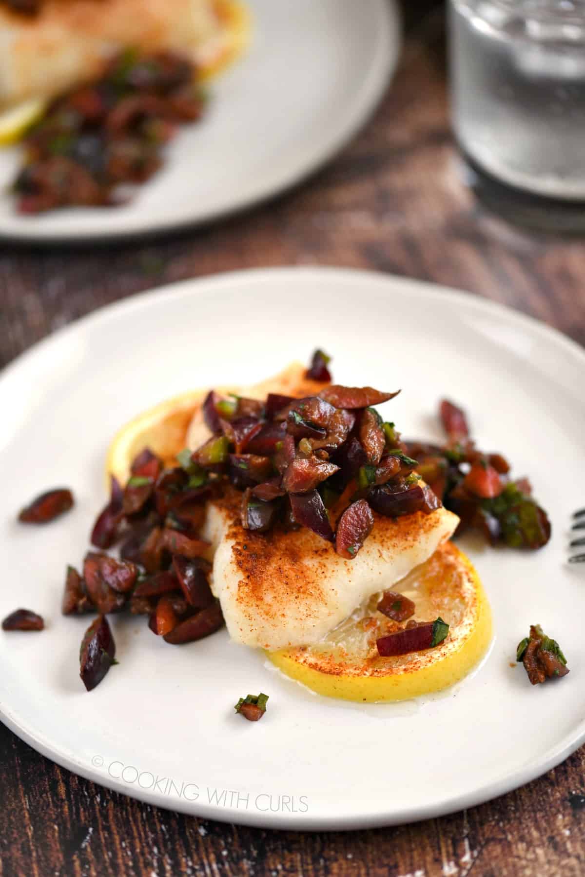 two white plates with lemon slices topped with baked cod and cherry salsa.
