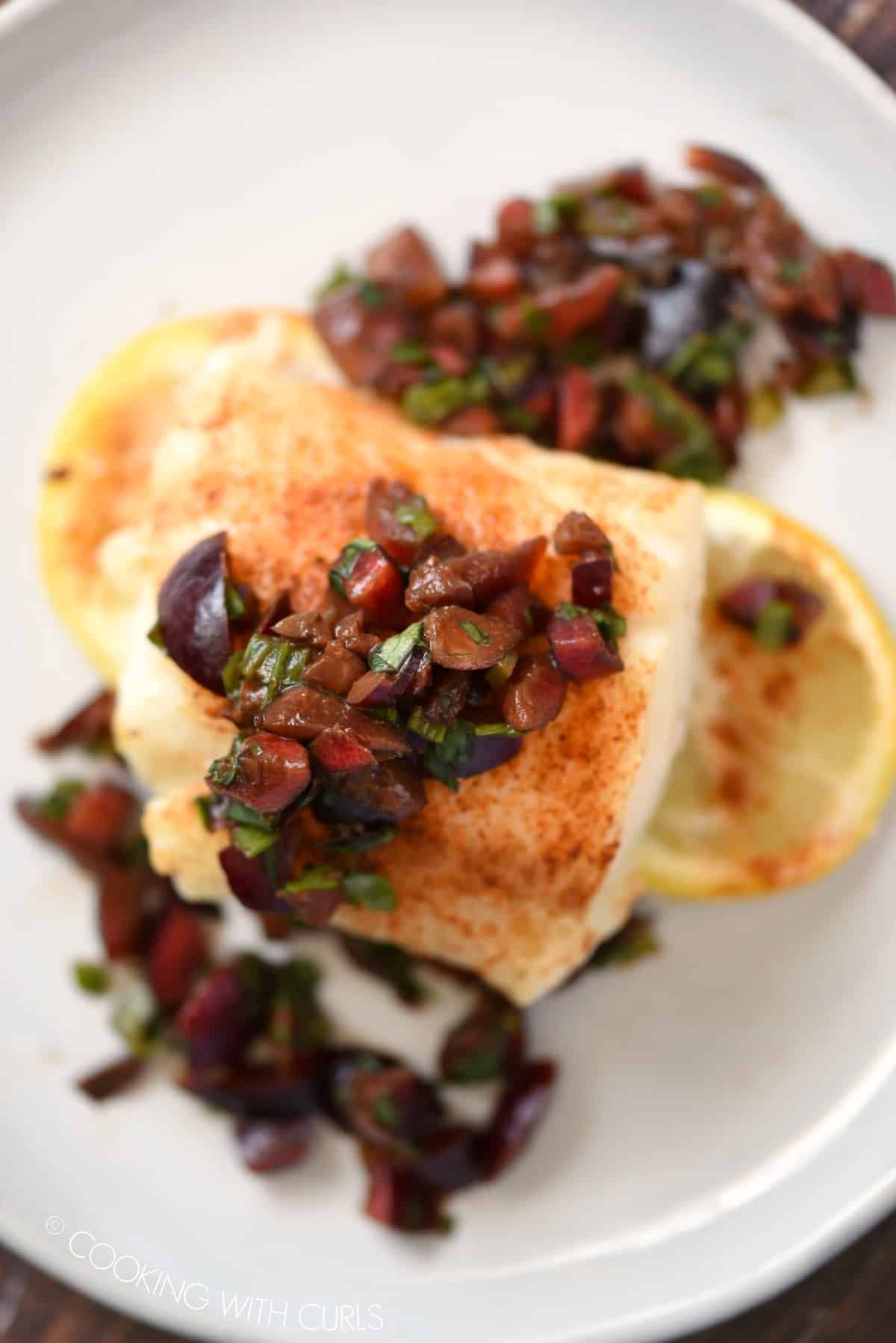 a close up of cherry salsa topped, baked cod sitting on two lemon slices on a white plate.