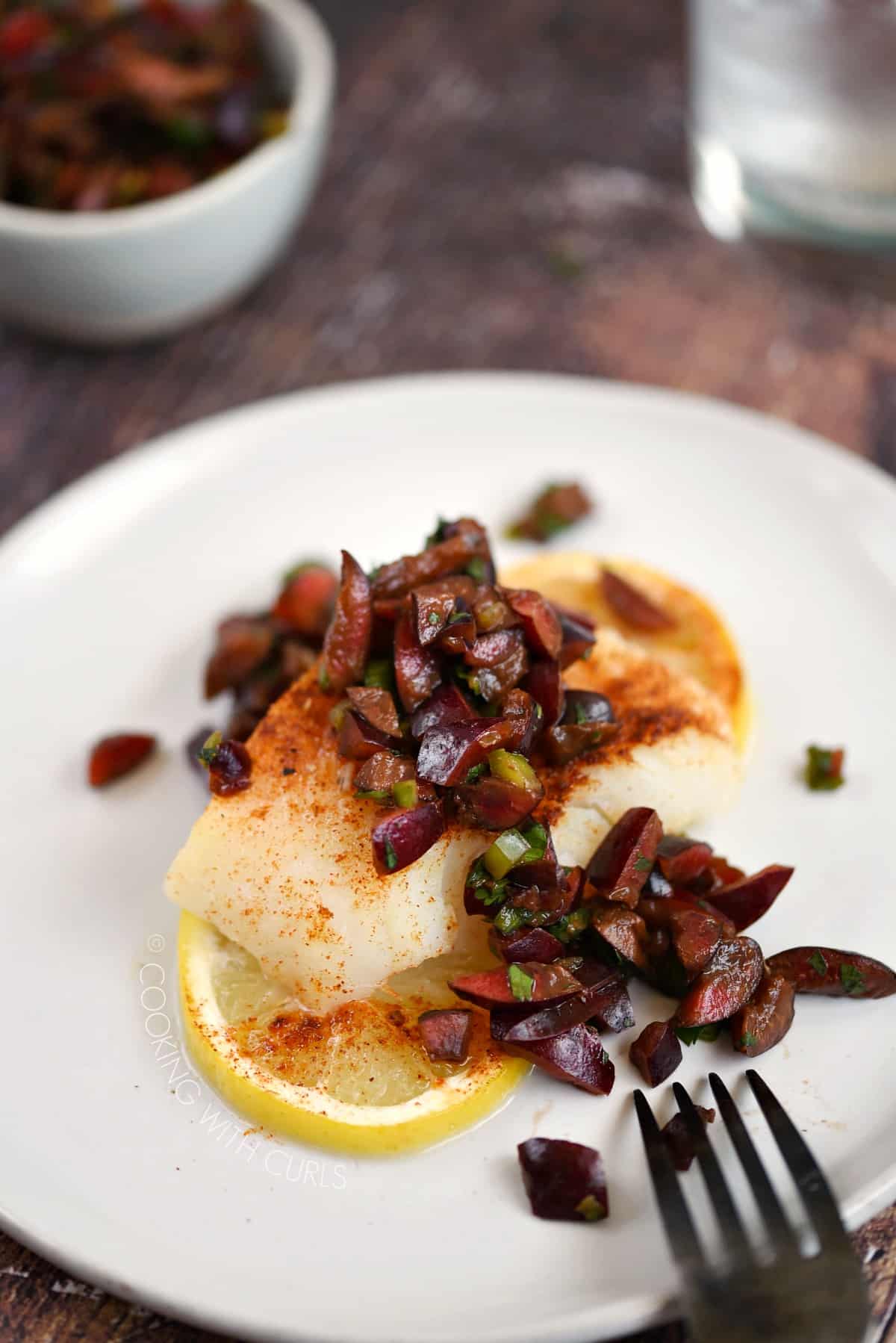 looking down on two lemon slices on a white plate topped with seasoned baked cod with cherry salsa on top and a bowl of cherry salsa in the background.