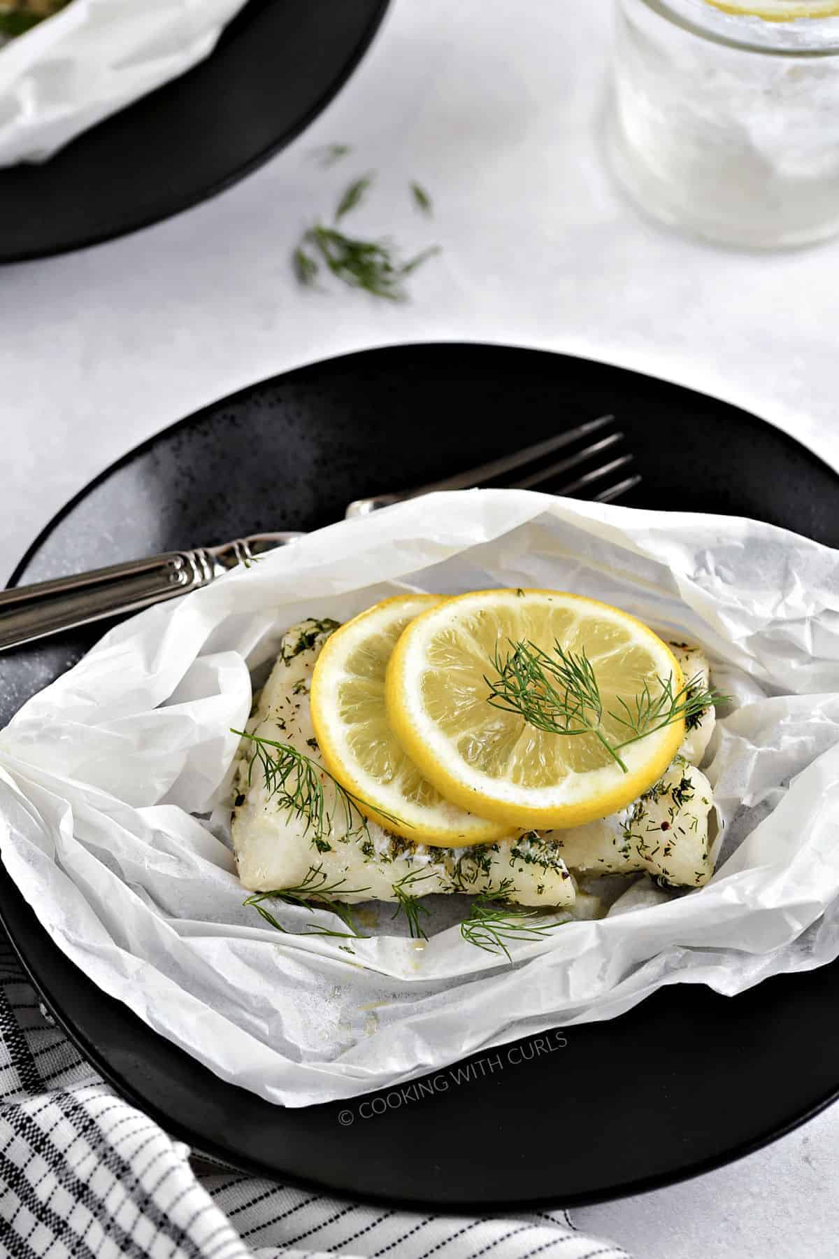 A cod filet topped with dill and a lemon slice in parchment paper sitting on a black plate.