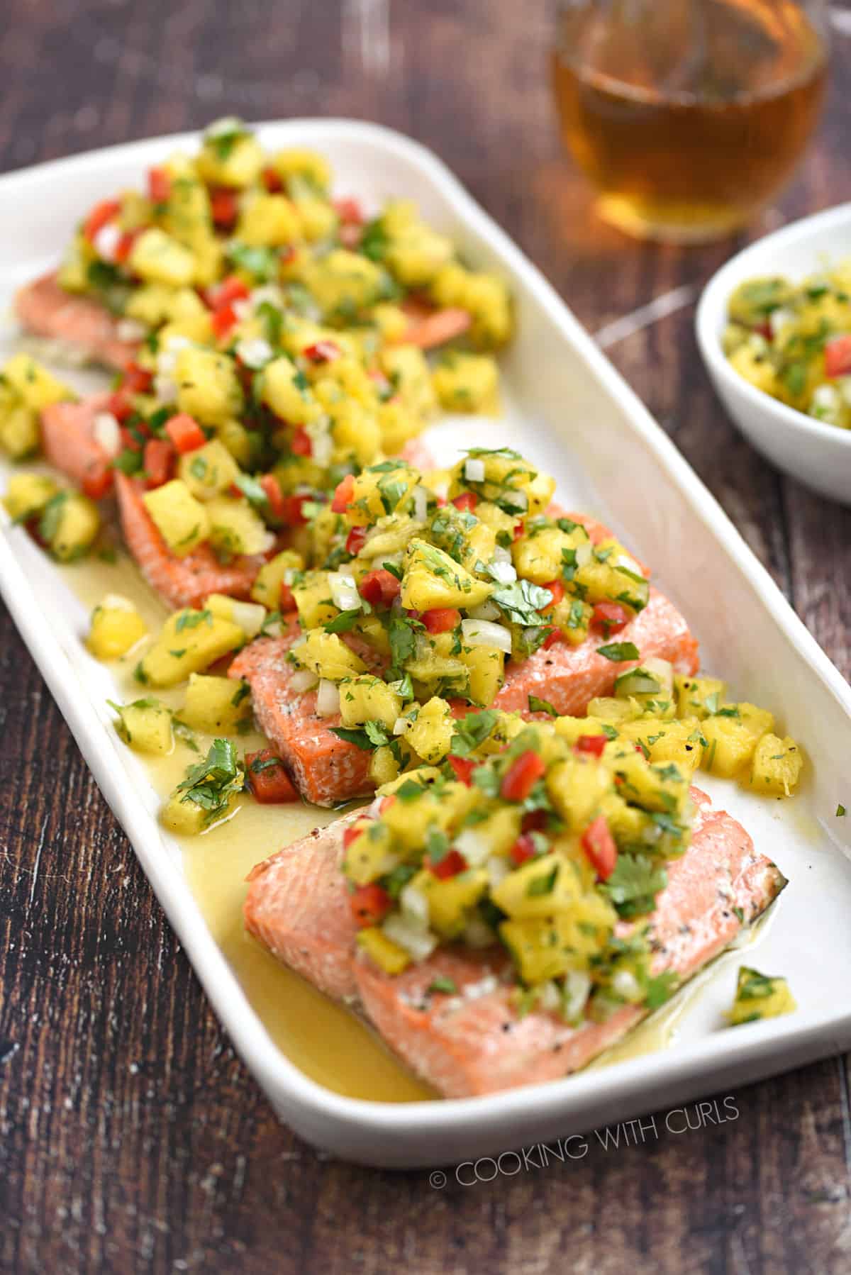 Four salmon filets topped with pineapple salsa on a white platter witha  glass of wine and bowl of salsa in the background.