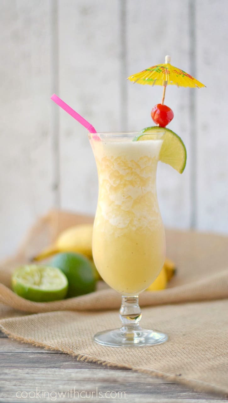 Looking at the side of a Frozen Banana Daiquiri in a hurricane glass garnished with a paper umbrella, lime wedge, and cherry.