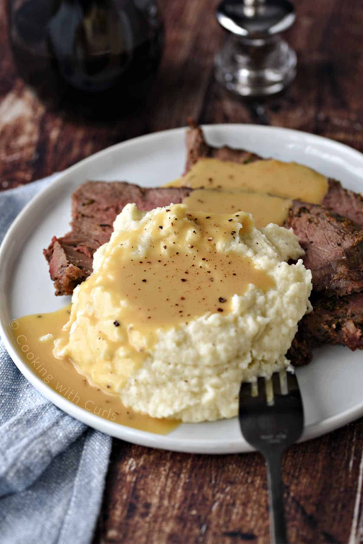 A plate of sliced roast beef and mashed cauliflower topped with beef gravy sprinkled with ground pepper.