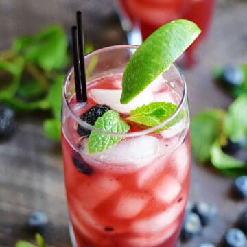 A pink drink in an ice filled glass with fresh blackberries, mint leaves and a lime wedge.