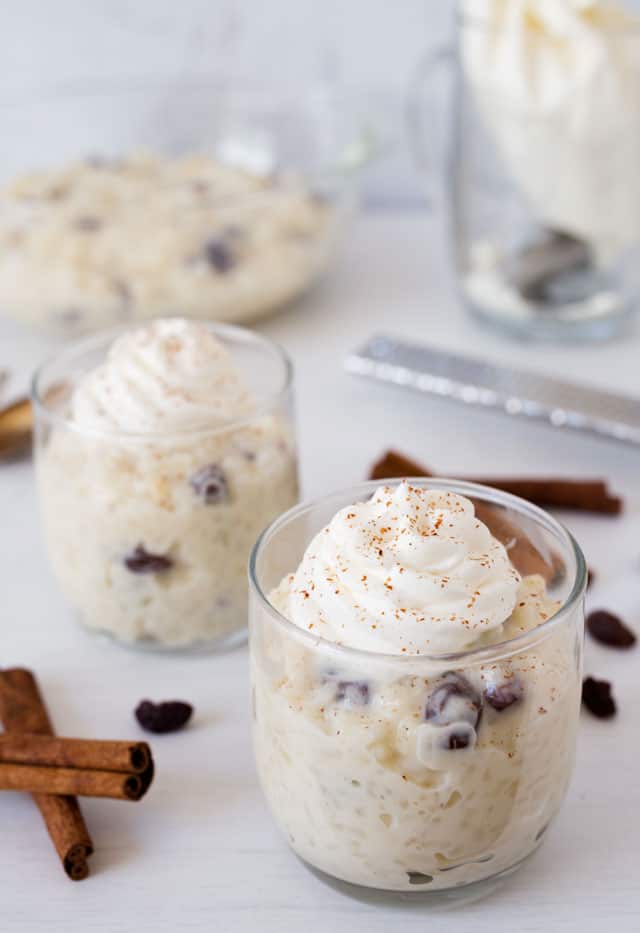 two small glasses of rice pudding topped with whipped cream surrounded by cinnamon stick, a grater and a bag of whipped cream in a glass jar.