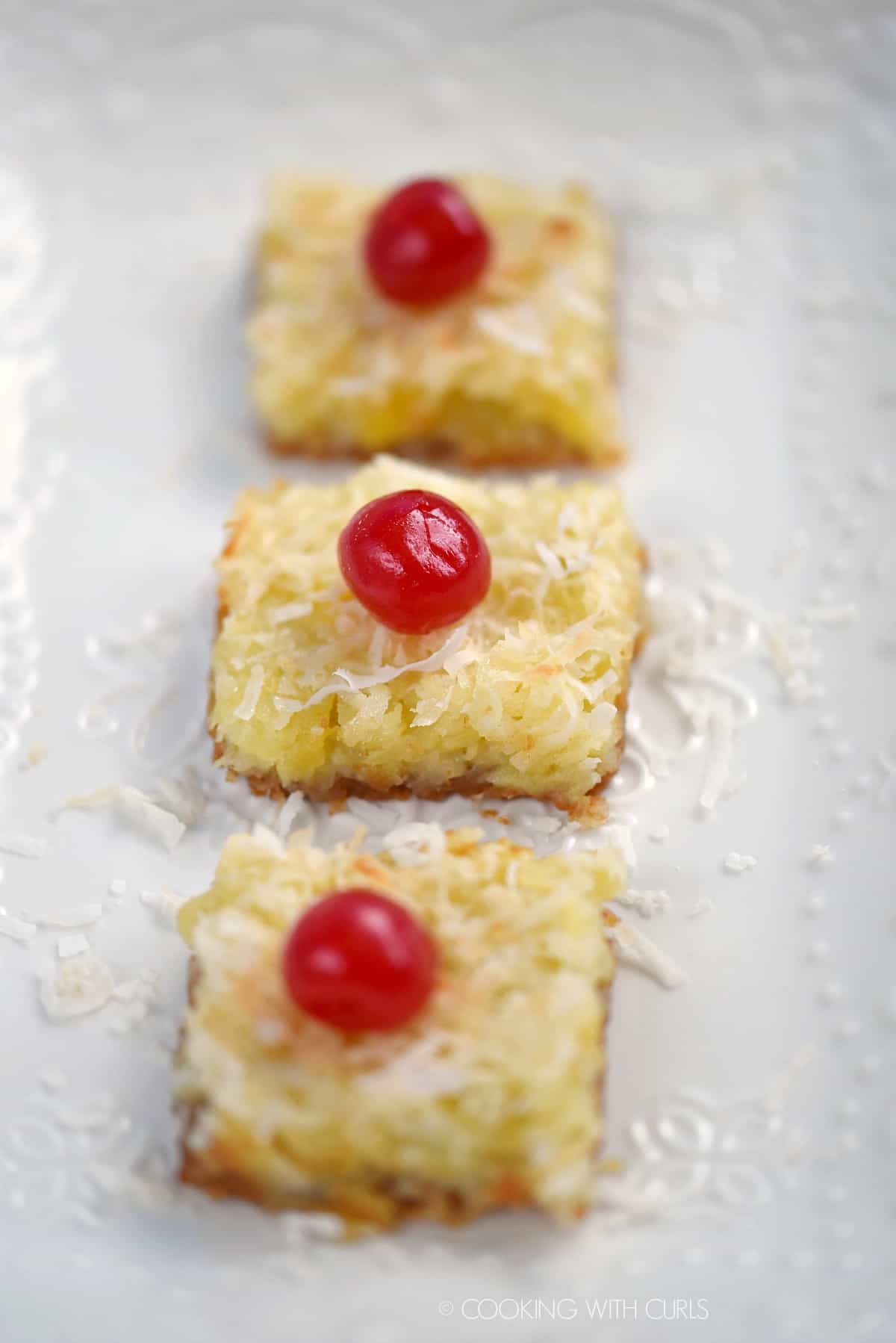 Three cherry topped Pina Colada Bars lined up on a white plate.