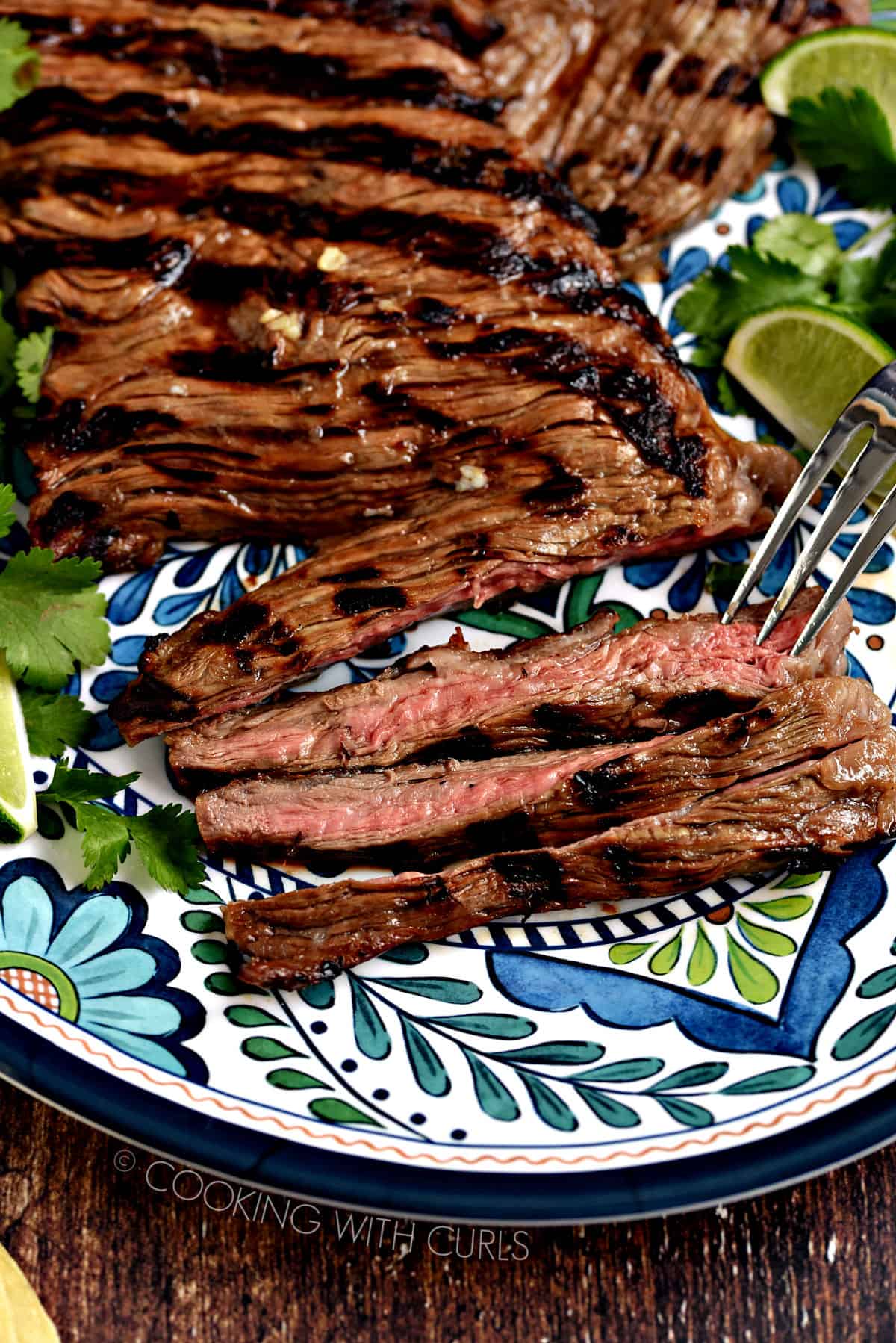 Grilled skirt steak on a decorative platter with lime wedges and cilantro leaves.
