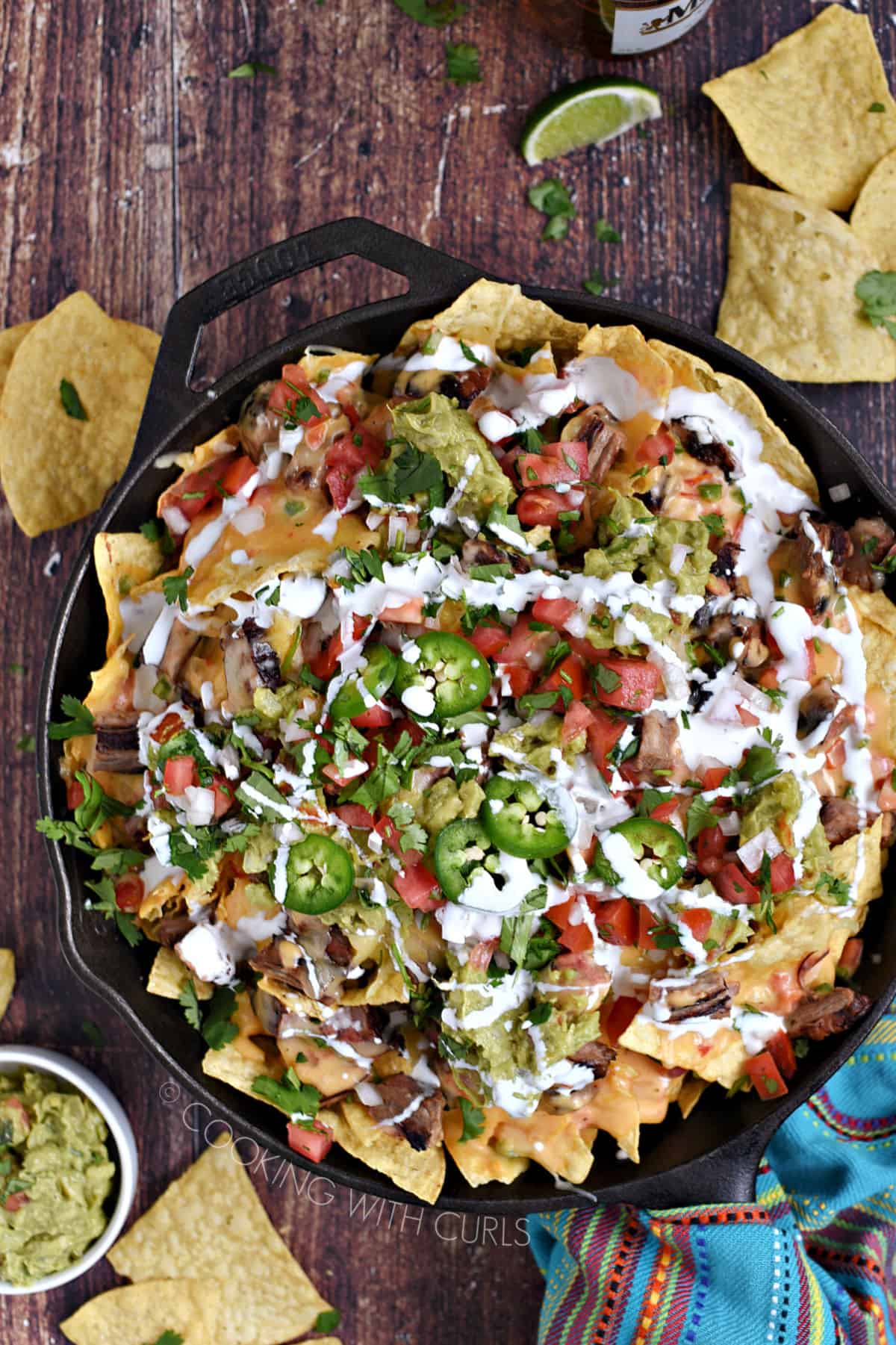 Looking down on Carne Asada Nachos in a cast iron skillet topped with crema, jalapeno and guacamole with chips scattered around and a bowl of guacamole in the background.