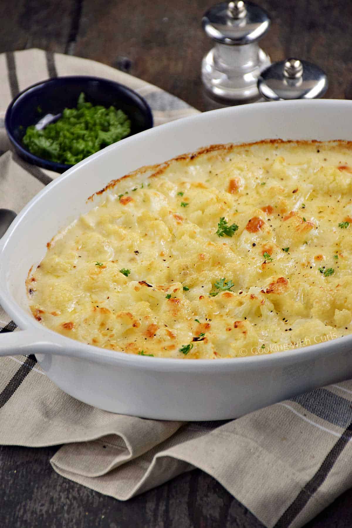 Cheese topped cauliflower in a creamy sauce in a white baking dish with salt and pepper shakers in the background.