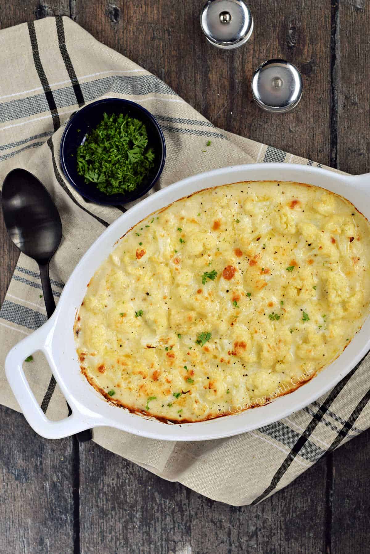 Cheese topped cauliflower in a creamy sauce in a baking dish with a small bowl of parsley in the background.