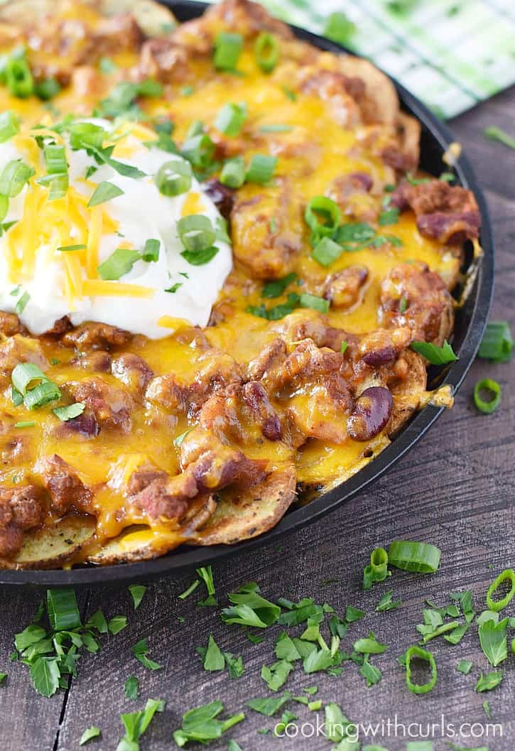 a cast iron skillet filled with potato slices topped with chili and cheese with sour cream in the center and sprinkled with green onions
