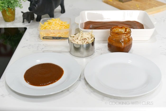 One plate with enchilada sauce and one empty plate on the counter with chicken and cheese in separate containers.