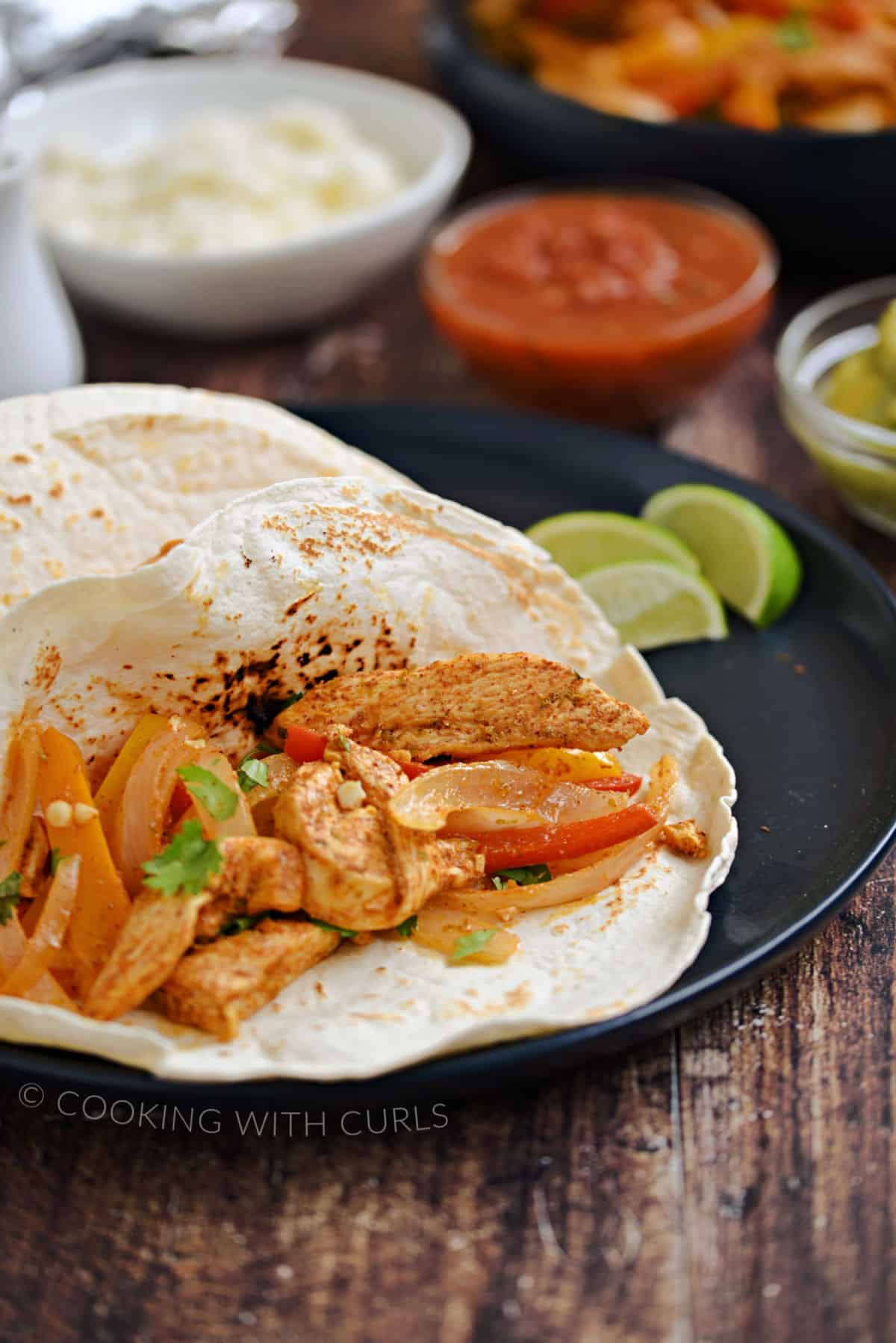 Chicken, onion, and bell pepper strips in flour tortillas with bowls of queso, salsa, and avocado in the background. 