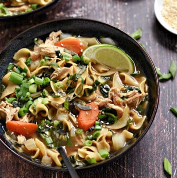 A bowl of Chinese Chicken Noodle Soup with carrots, green onions, and lime wedges.