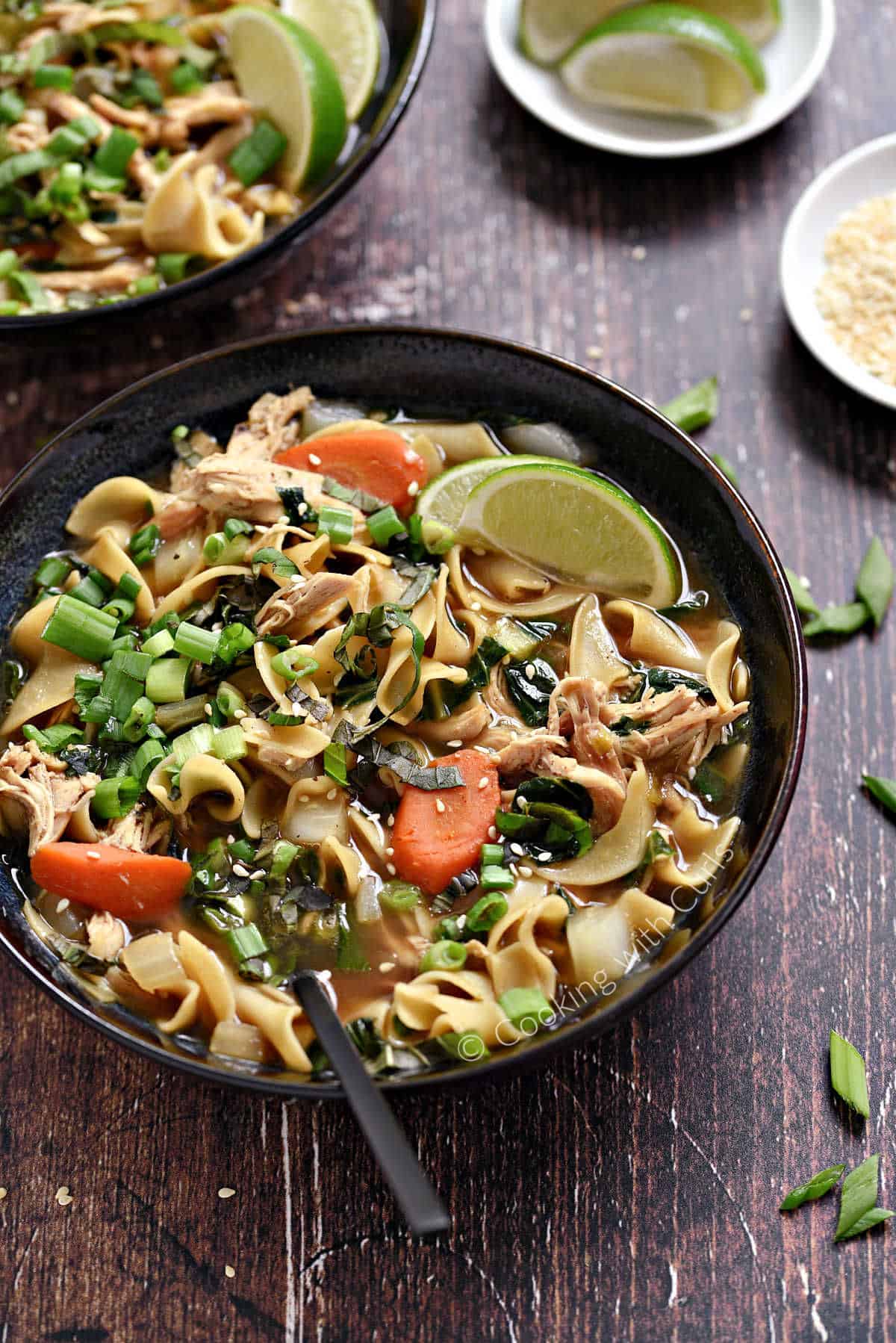 A bowl of Chinese Chicken Noodle Soup with carrots, green onions, and lime wedges. 